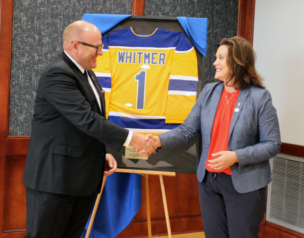 LSSU President Hanley and Governor Whitmer shake hands as she accepts a personalized jersey from LSSU